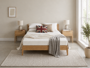 A light oak wooden bed frame with a white textured mattress, styled with white pillows, a Union Jack accent pillow, and a gray throw blanket, set in a bright, modern bedroom.