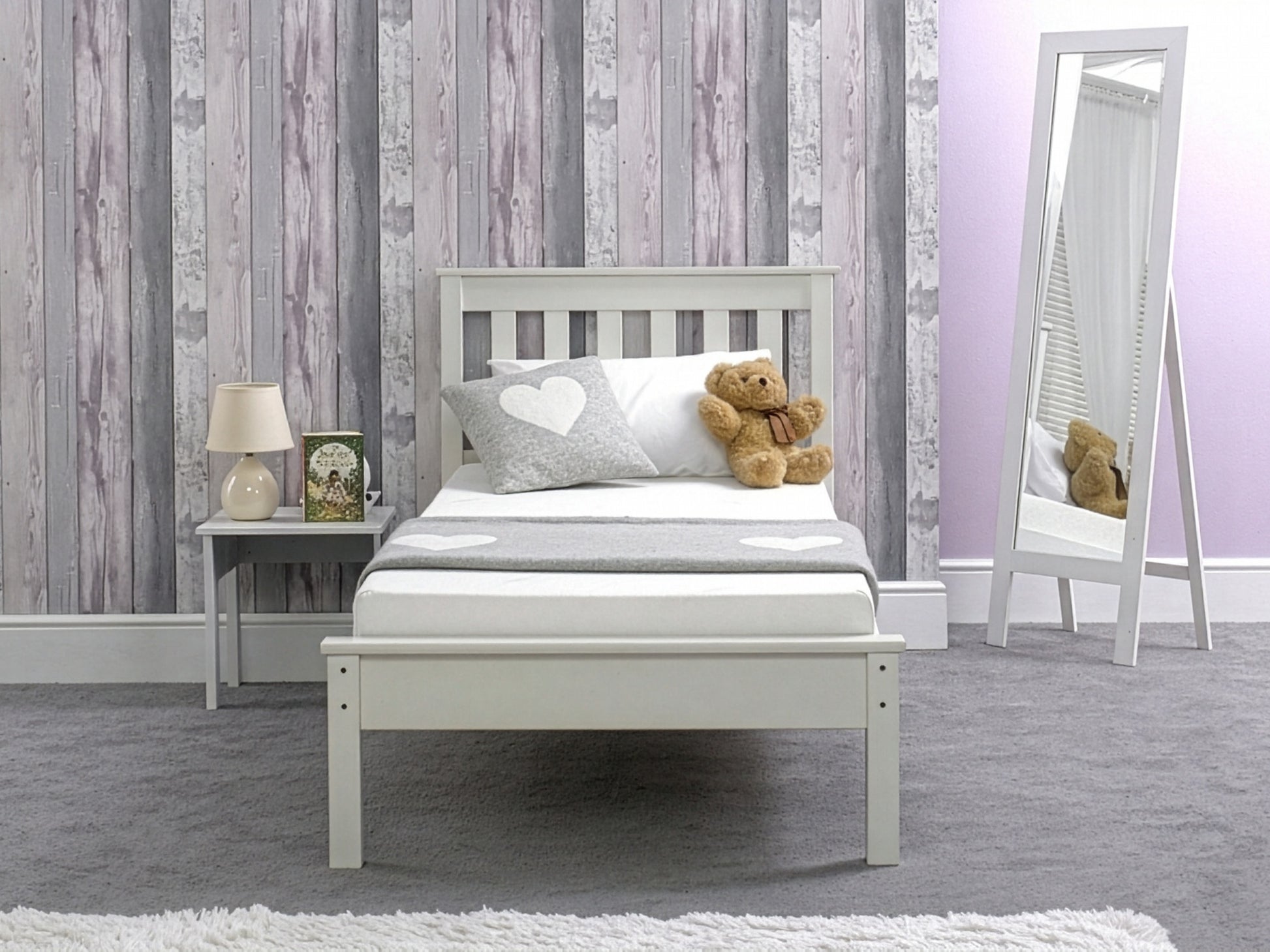 A child's bedroom featuring a white bed frame with a memory foam mattress, a grey rug, and a teddy bear on the bed. The wall behind the bed features a wood panel design in grey tones.