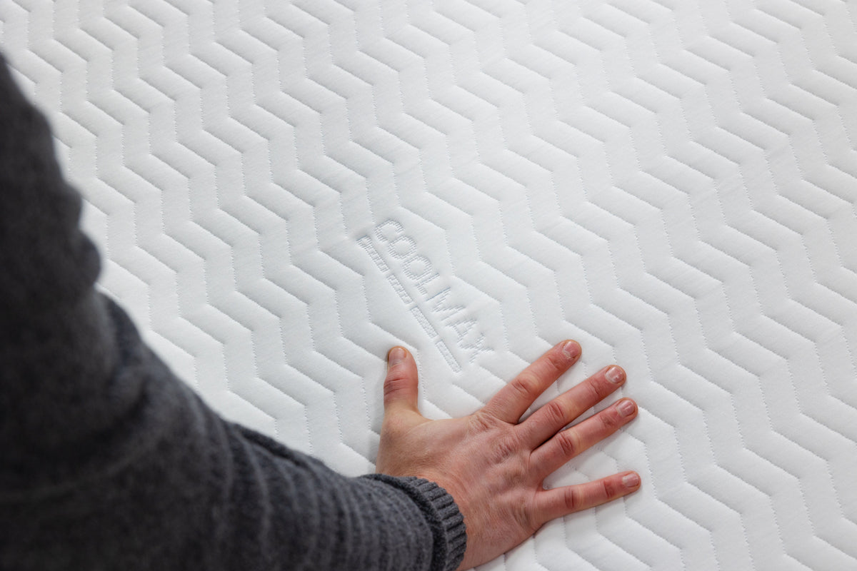 A close-up of a white mattress topper or mattress surface with a textured chevron pattern, showing the embossed brand name 'COOLMAX'. A person's hand and arm are resting on the surface to demonstrate its texture and comfort.